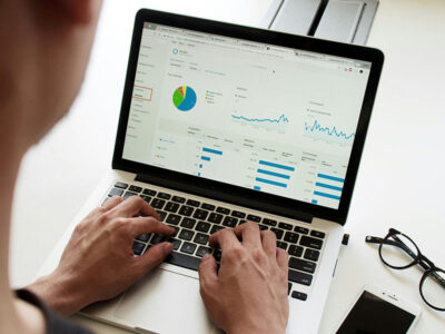 The image is taken over the shoulder of person sitting in front of their laptop. The screen shows many different info and data charts.