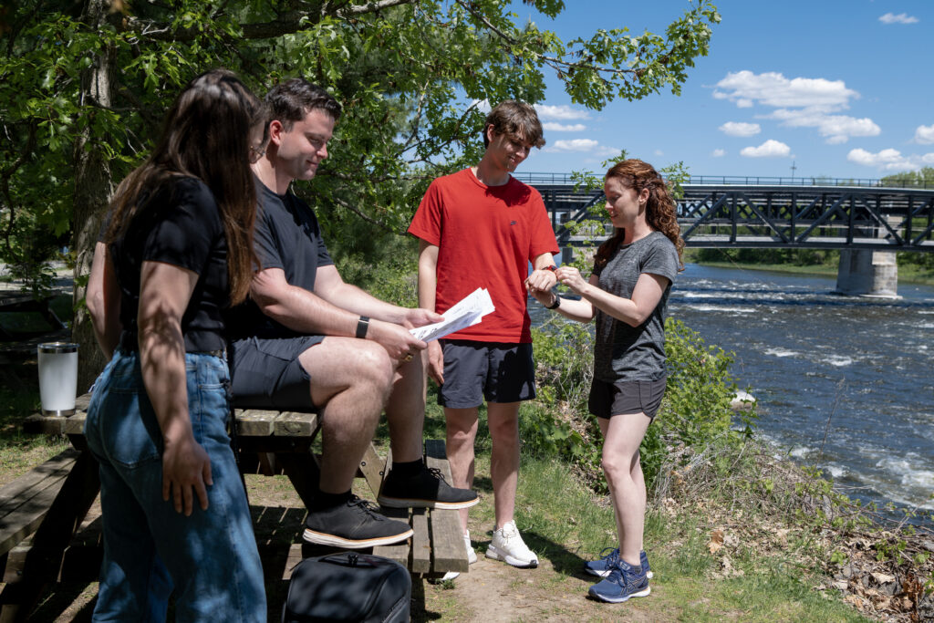 Four people sit and stand in front of river looking at accelerometers and data