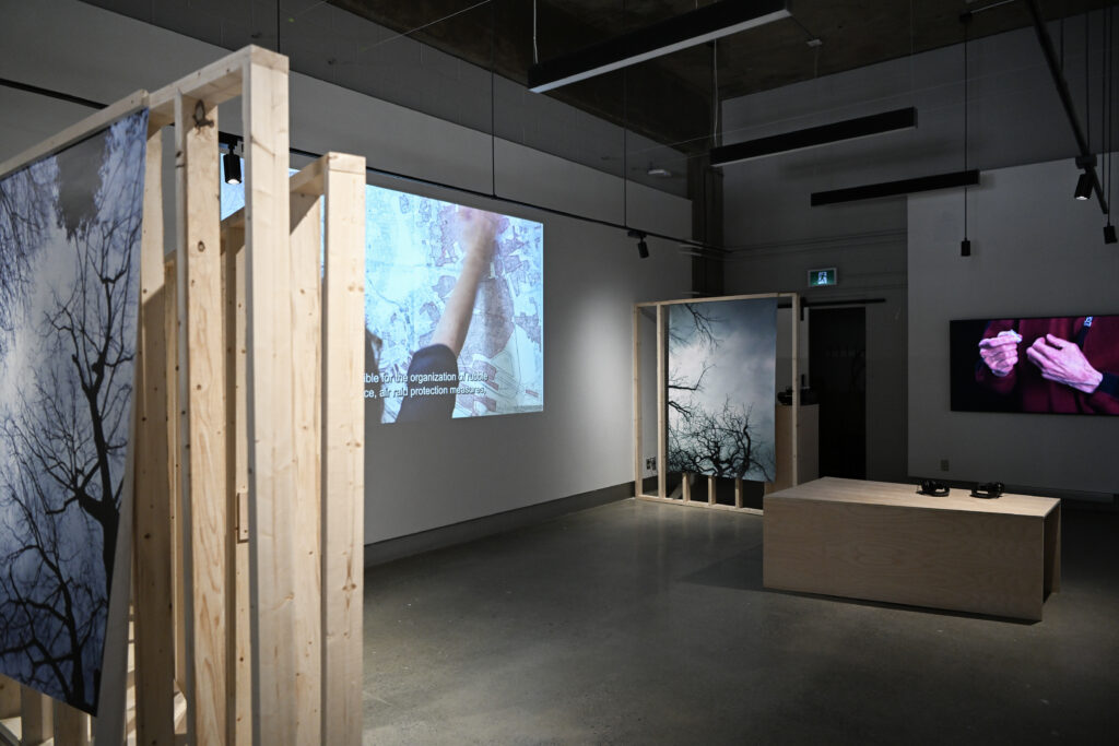Photograph of exhibition showing two video projections of maps and ruined objects as well two photographs of leafless trees.