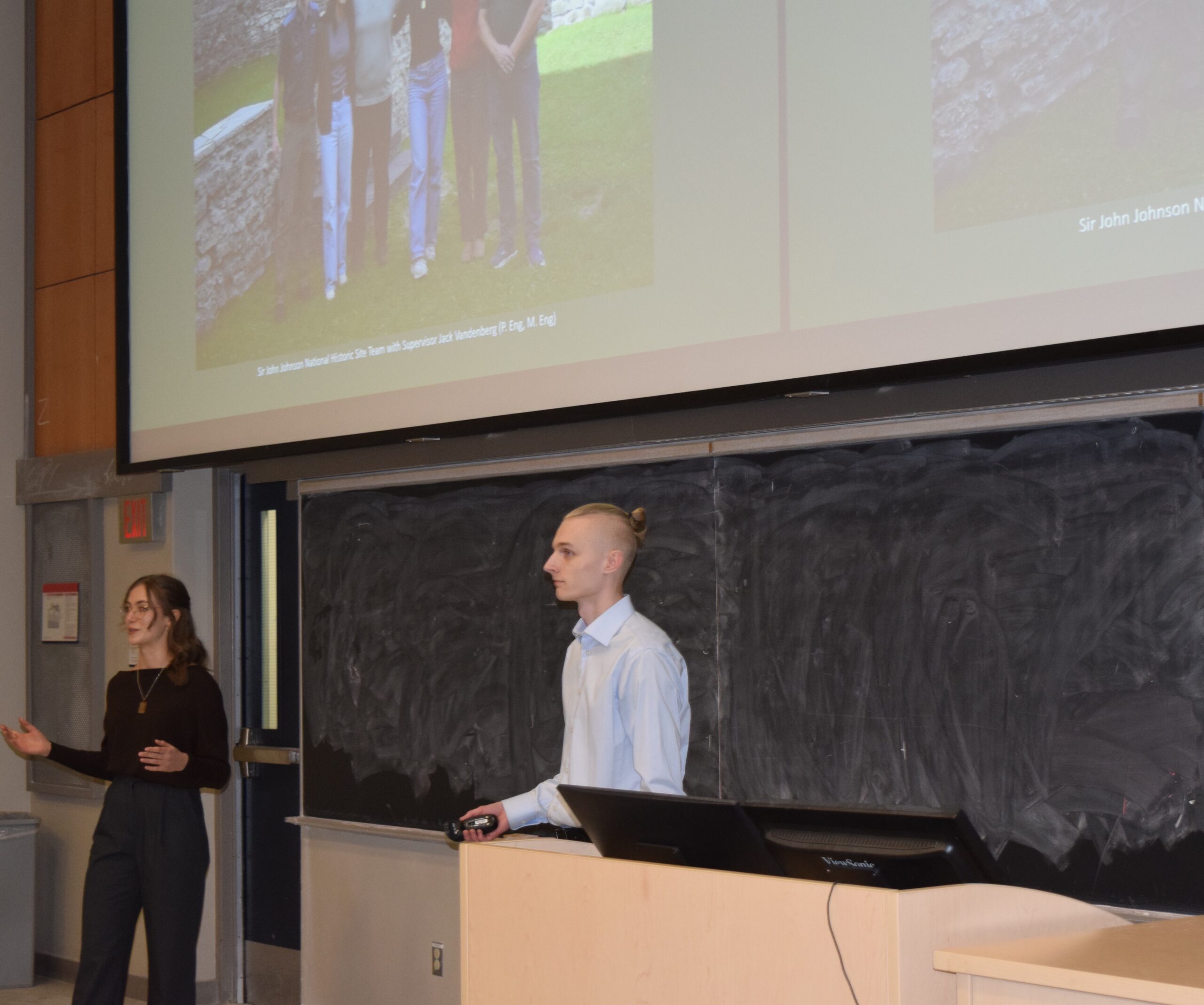 Another angle of Maclean Crowdis and Spencer Watson presenting their project in front of a lecture hall in formal clothing.