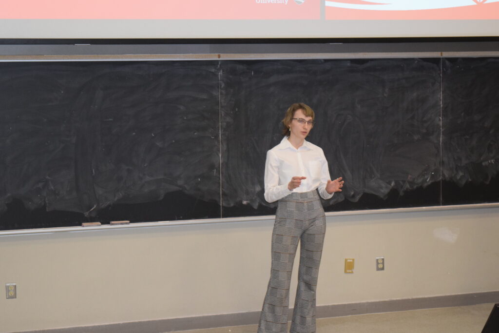 Photo shows Sophie Cormier presenting their group's capstone project in front of a lecture hall in formal clothing.