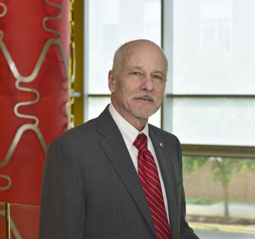 Image shows Dr. Jim Milke wearing a grey tuxedo with a red tie and a small pin on his left lapel.
