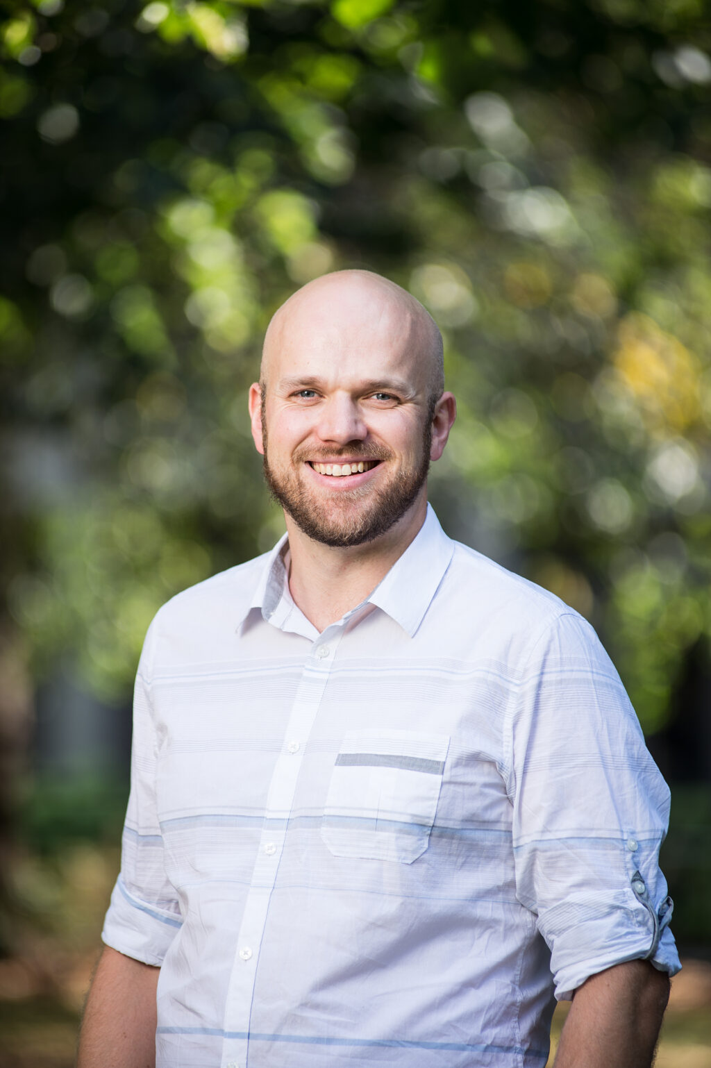 Photo of smiling Liam O'Brien wearing blue shirt in front of a greenery.