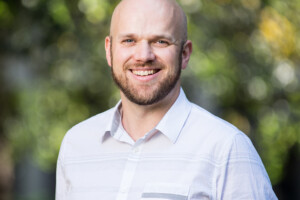 Photo of smiling Liam O'Brien wearing blue shirt in front of a greenery.