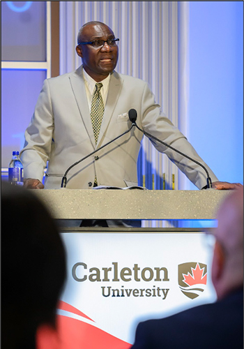 A man in a light grey suit delivers a speech from a podium about AI in Canada.