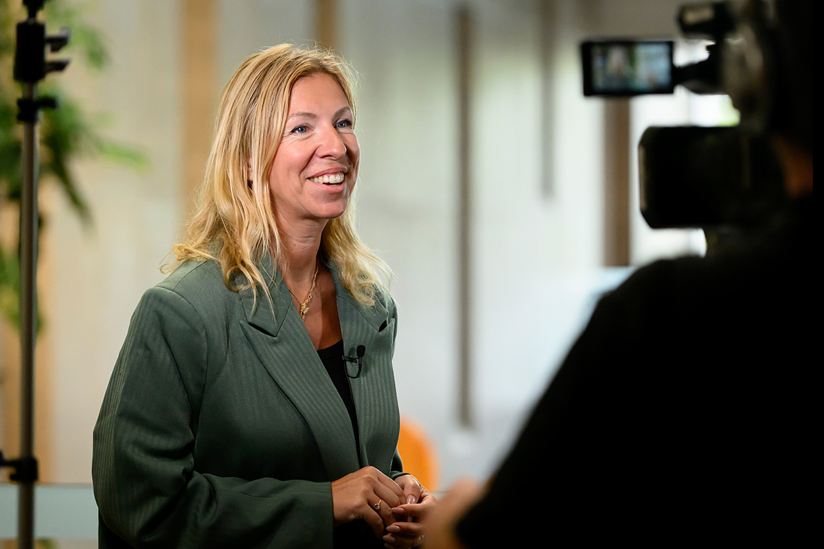 A woman wearing a green blazer speaks into a camera.