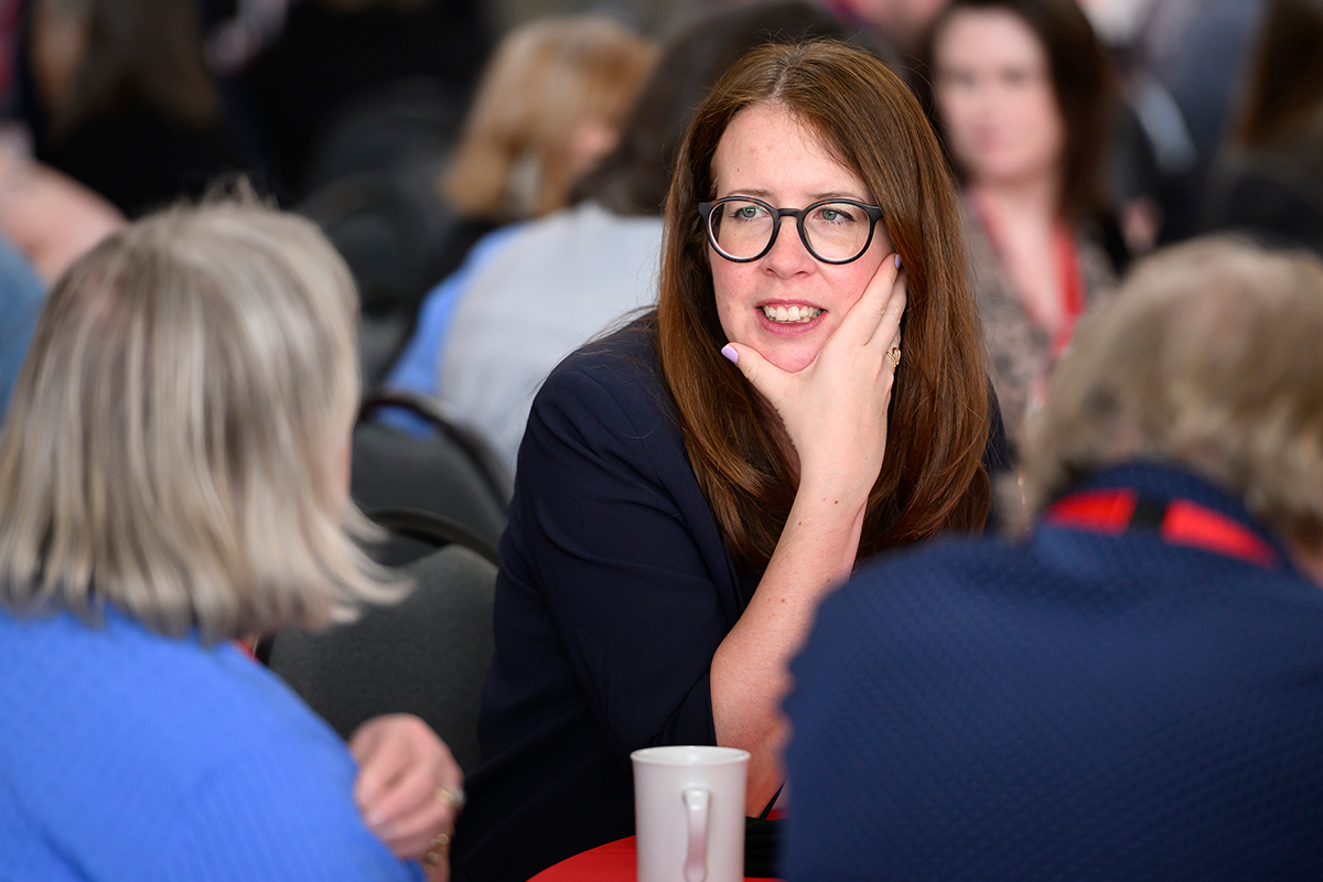A woman wearing glasses holds her hand to her face while engaging in conversation.