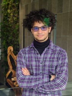 2022 Undergraduate Co-op Student of the Year, Abby Ibrahim, poses with his arms crossed in front of a tiled wall with a green leafy plant in the background.