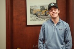 2022 Undergraduate Co-op Student of the Year Austin Herrrington wears a Cavanagh baseball hat and stands in front of a brown wall in front of a photograph of a yellow construction truck.
