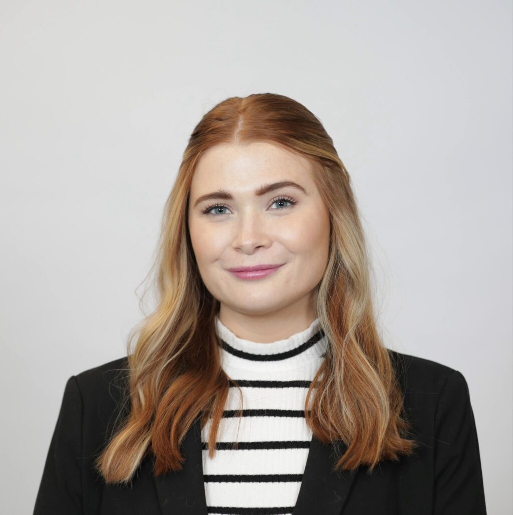 Savanna Platt stands against a grey wall wearing a black and white striped shirt and black blazer.