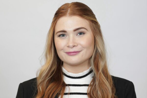 Savanna Platt stands against a grey wall wearing a white and black striped shirt and black blazer.