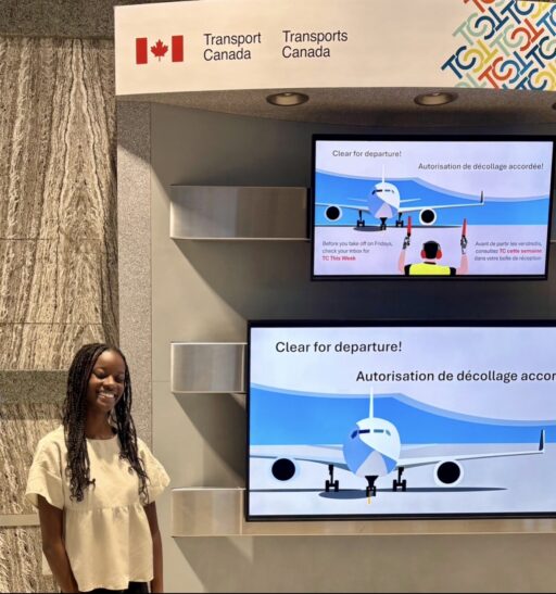 Makayla stands in a Transport Canada building. There are TV screens with aviation images next to her.