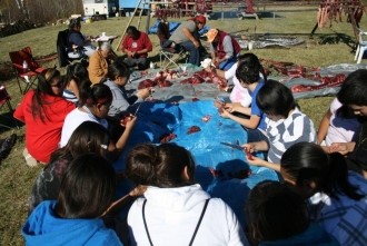 Youth sitting around a blue tarp on the ground, learning how to make dry meat.