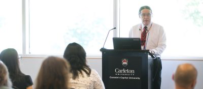 Professor John Medicine Horse Kelly stands at a Carleton University podium, speaking to an audience.