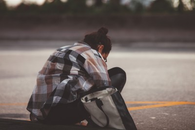 A young woman sits on a curb with her head down and her bag beside her.