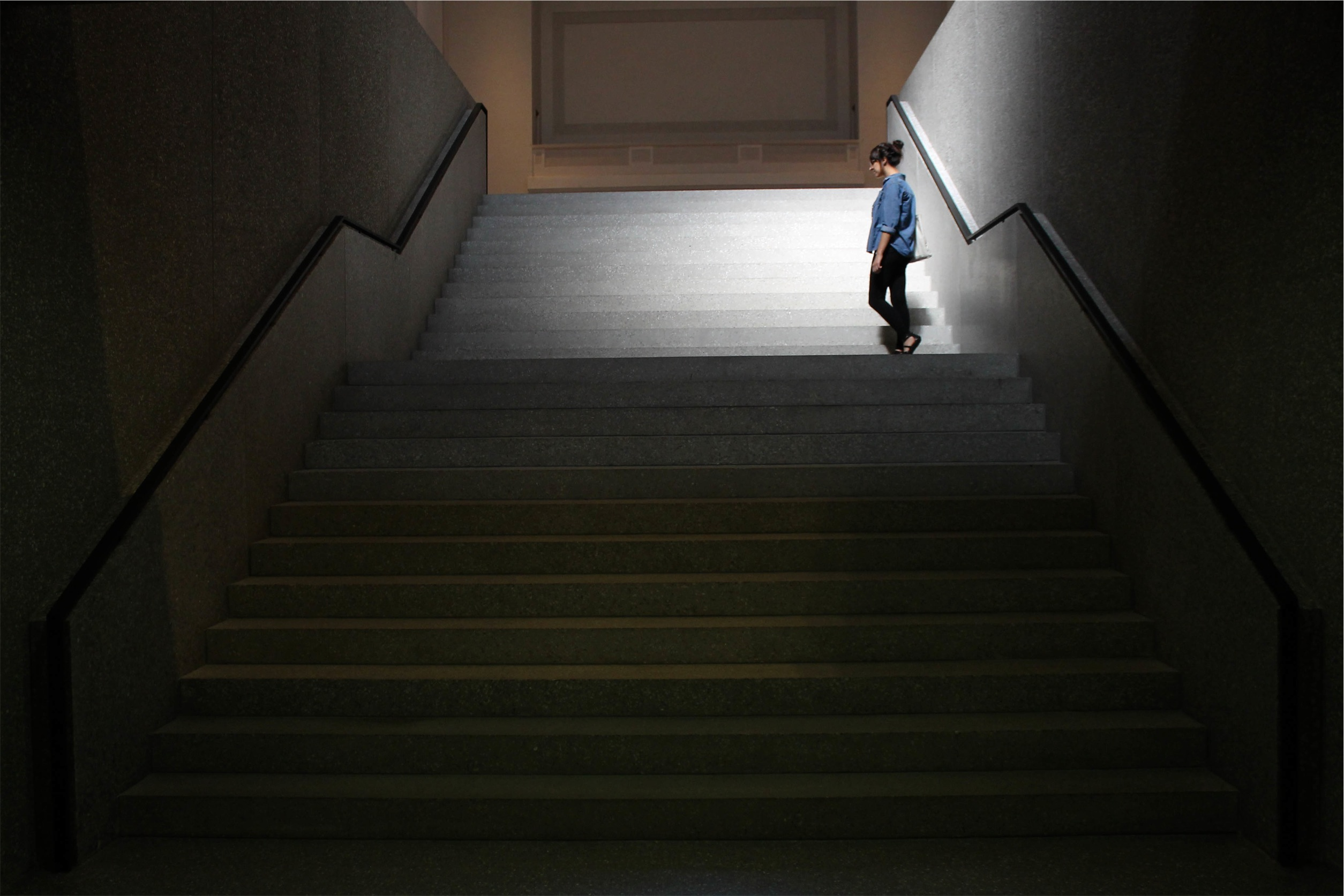 A young woman waits at the top of a long dark staircase.