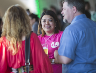 A female volunteer in a pink C2UExpo 2015 shirt smiles at two conference delegates whose backs are turned to the camera.
