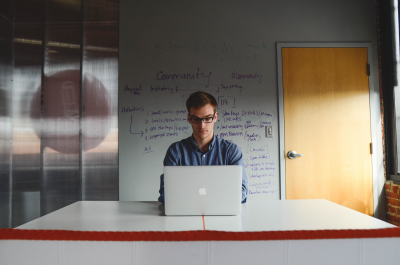A man sits at his apple laptop in front of a white board covered in start-up key words such as "community".