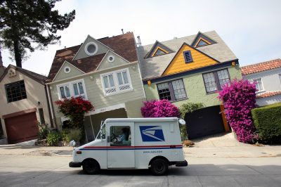 A picture of a mail truck parked on a hill with houses in the background. The picture is taken with the car aligned horizontally instead of the houses so the houses look as if they are slanted to the left.