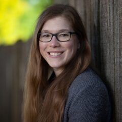 This is a headshot of a woman is leaning against a brown wall looking at the camera.