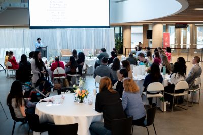 Photo of past conference's participants sitting at round tables, facing a presentation.