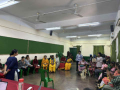Looking from the front, a facilitator leads the workshop, using a presentation slide and whiteboard for discussion with participants.