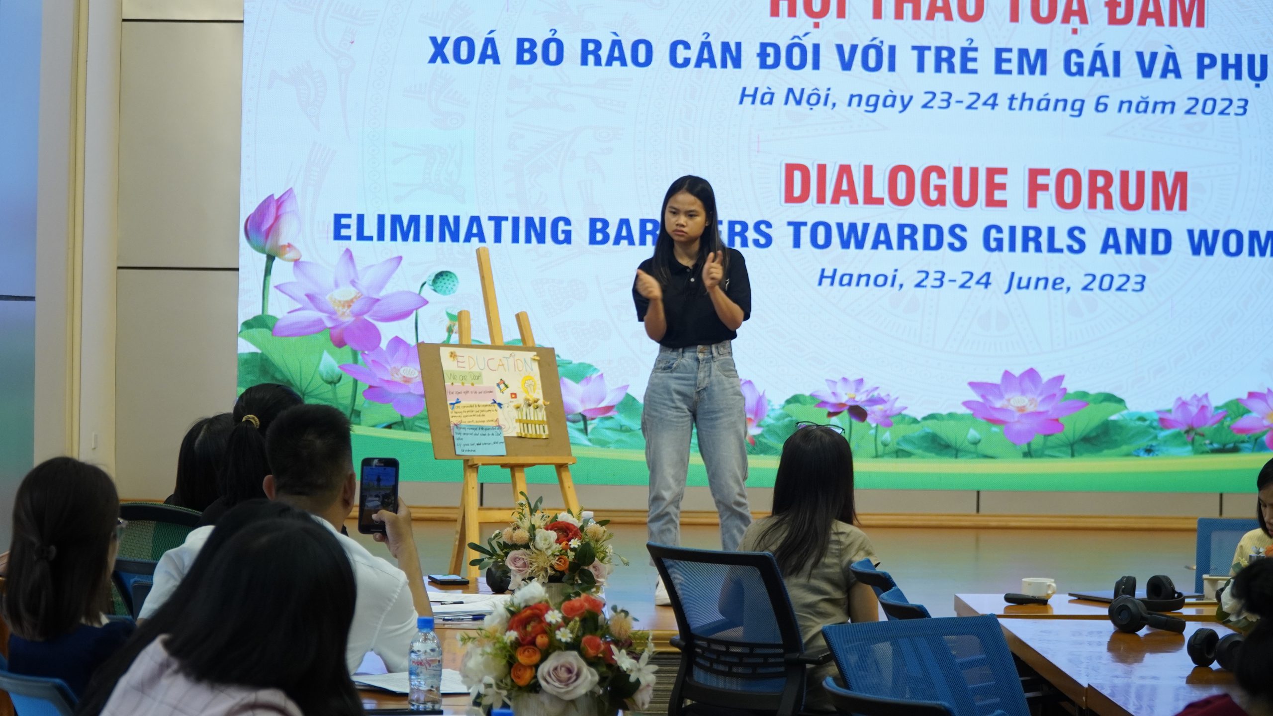 There is a girl wearing a black T-shirt and jeans presenting her manifesto using sign language, and the drawing is placed next to her.