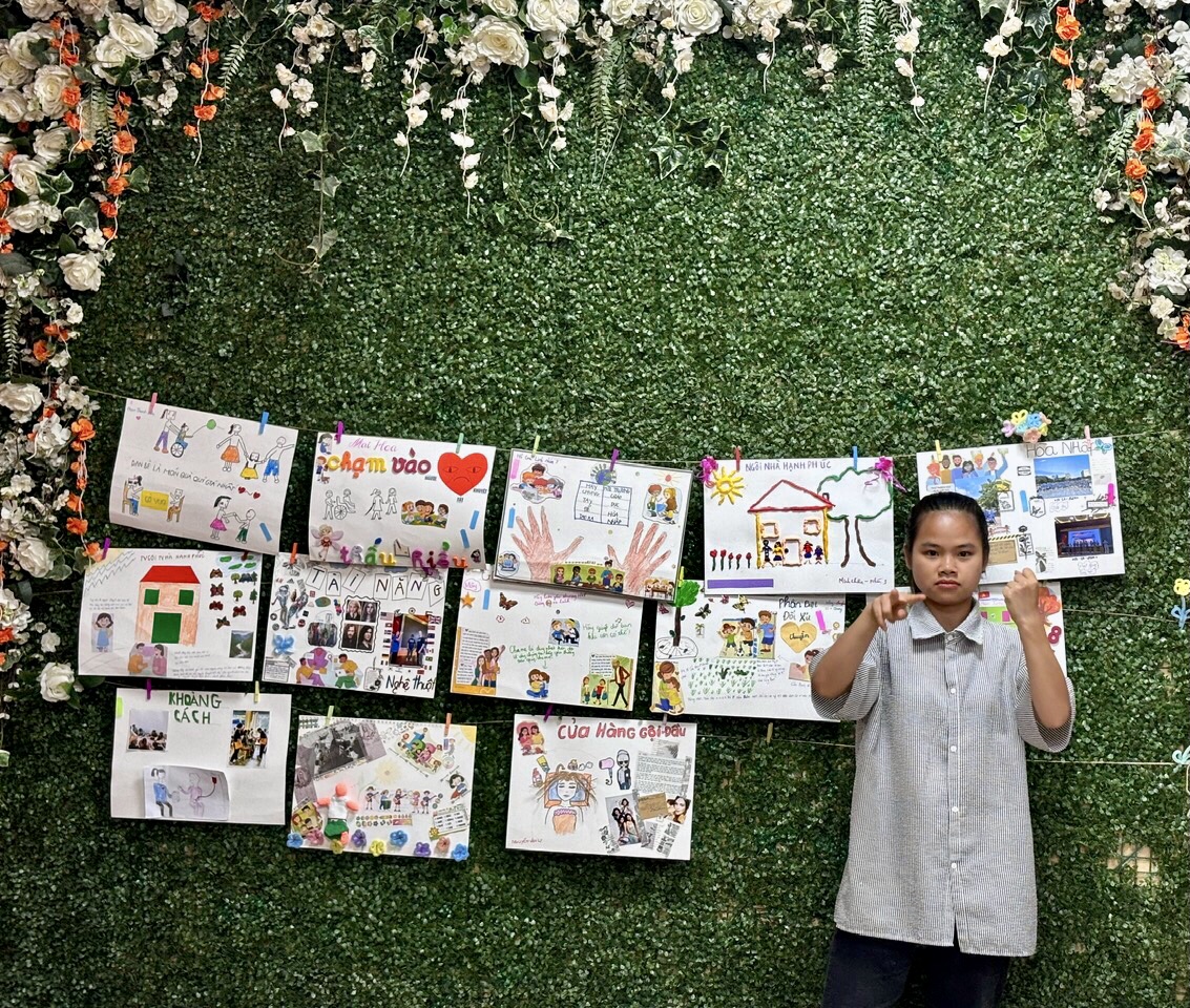 A girl is presenting using sign language. Behind her is a green background with various drawings hung up.