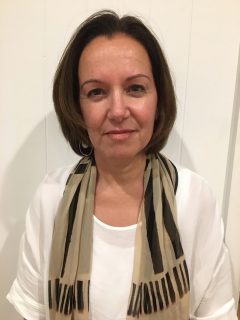 This is a photo of a woman with short brown hair. She is wearing a white shirt and a brown patterned scarf.
