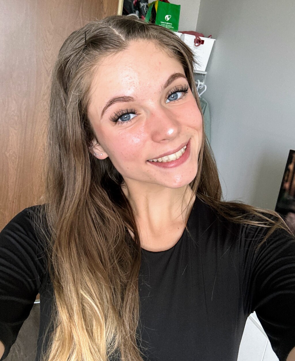 A smiling person with long light brown hair wearing a black top, looking at the camera in a well-lit indoor space.