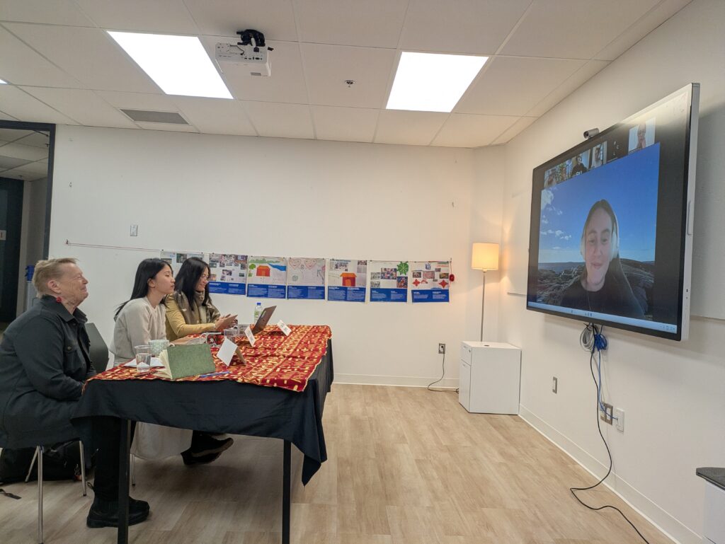 Three people sitting at a table with a Zoom screen showing the facilitator and online participants.