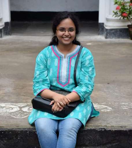 A person wearing a teal Kurt and glasses, seated outdoors on steps with a bag on their lap, smiling at the camera