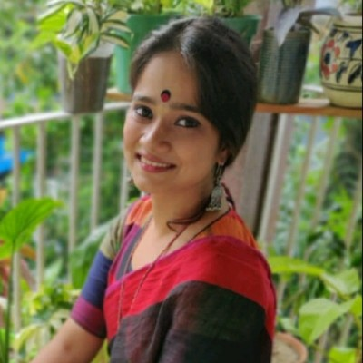 A person wearing a red and black sari with a bindi, seated on a balcony surrounded by green plants, smiling at the camera.
