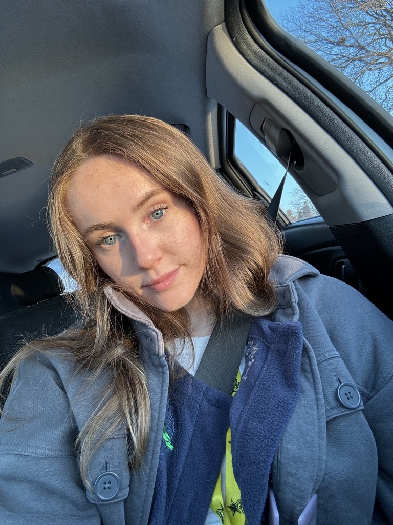 A person with shoulder-length brown hair wearing a grey coat, sitting in a car with sunlight illuminating their face