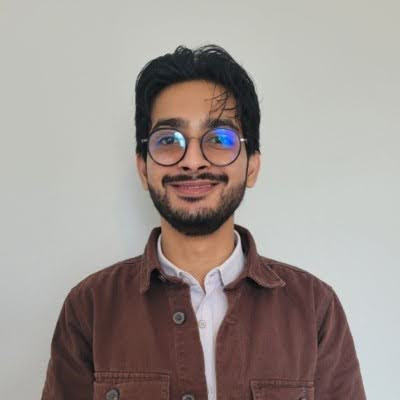 A person wearing glasses and a brown jacket over a white shirt, smiling in a portrait against a plain background
