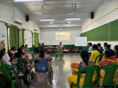 Looking from the back, a facilitator leads the workshop, using a presentation slide and whiteboard for discussion with participants.