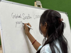 A person with long dark hair tied back is writing on a whiteboard with a black marker. The text on the whiteboard is "Global South CONNECT" and "young". The person is wearing a light blue shirt and has a red bangle on their right wrist.