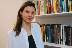 This is a photo of a woman with long brown hair wearing a black and white outfit. Behind her is a bookshelf.