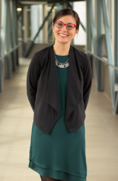 This is a photo of a woman wearing an emerald green dress, with a black vest on top. She has brown hair and red glasses.