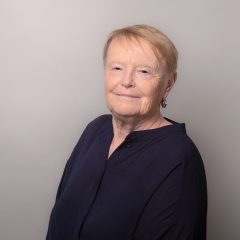 This is a portrait of a woman who has short blond hair, and who is wearing a dark blue shirt. The background is gray.