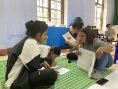 There are a girl and a woman sitting on a mat placed on the ground. The woman is holding a zine. Behind them, there is a girl writing and another girl holding her group's zine