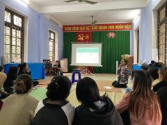 A woman with disabilities is giving a presentation. Behind her is a projector. Other participants are sitting in a U-shaped circle.