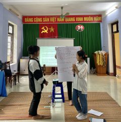 Two girls are holding mics and having a presentation. Behind them is a woman holding a poster