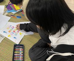 : A girl sitting on the ground is coloring a zine titled “Community inclusion"