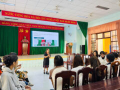 A woman with disabilities presents in front of a group of people. A large screen on stage displays a presentation titled “ENGAGE: Reporting research results with community 2022–2025.