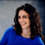 Women with dark brown curly hair smiling wearing a blue sweater. 