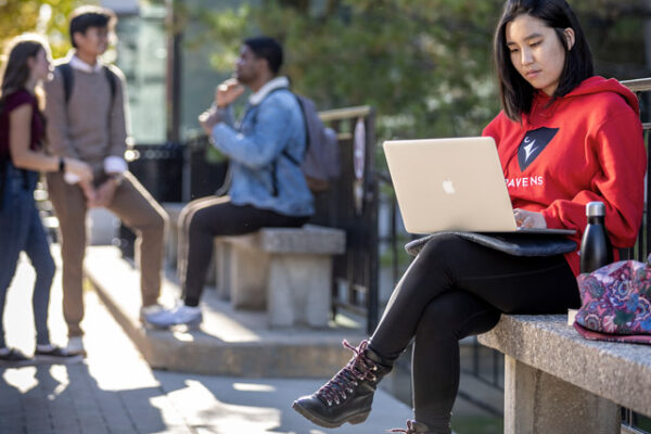 A student sits outside on a bench in Carleton's Quad using a laptop while other students talk in the background.