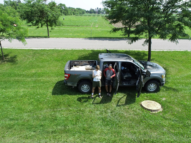An aerial view of two people standing next to a car, looking up at the camera on a drone.