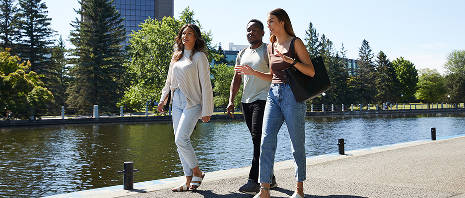 Three students walking along a riverbank with a tall building in the background