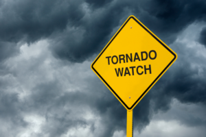 Diamond shaped yellow sign with black text reading tornado watch with stormy clouds in background.